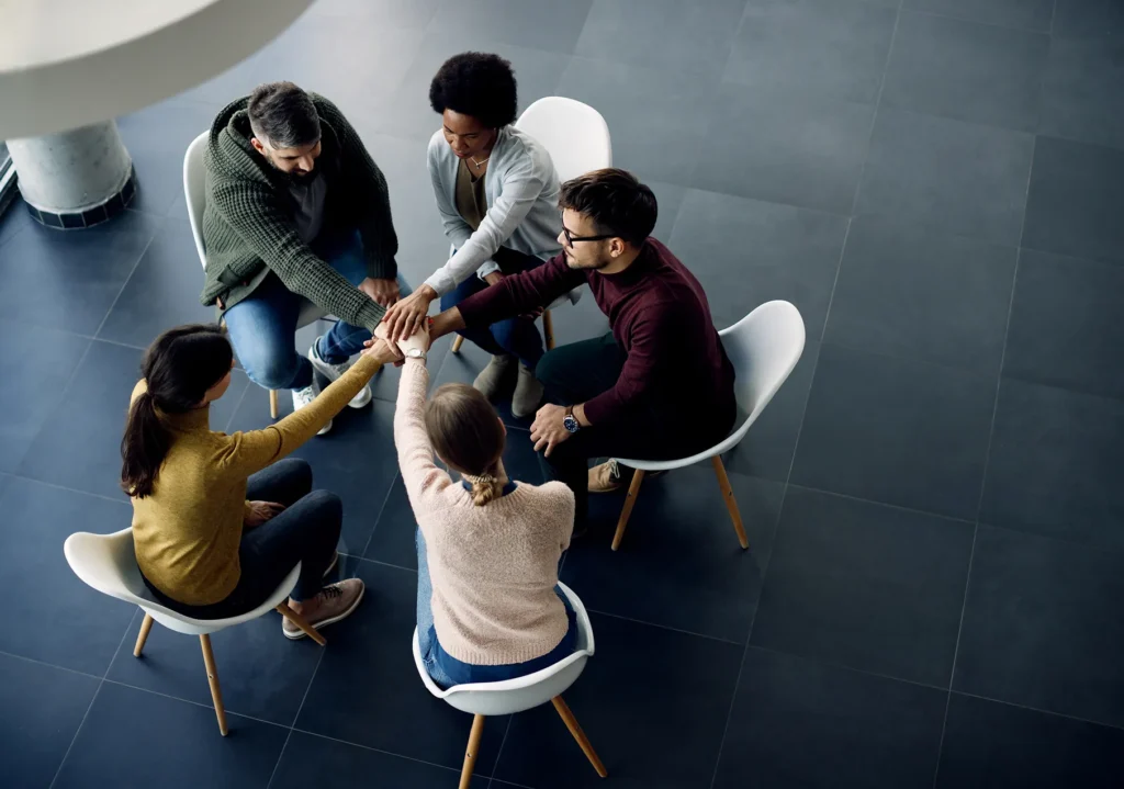 A diverse group of five people sitting in a circle, placing their hands together in the center to symbolize collaboration and successful conflict resolution through mediation.