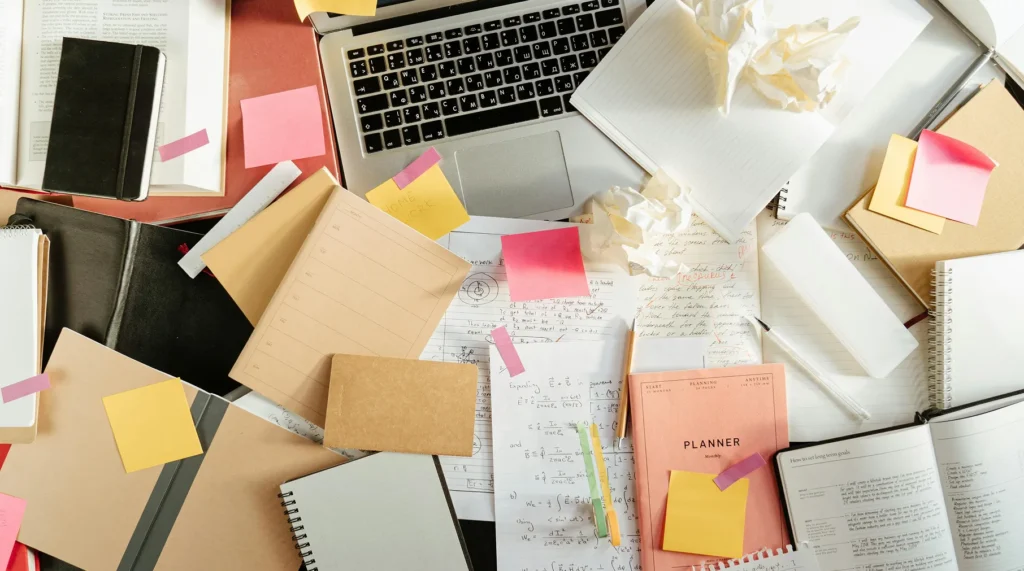An overhead view of a cluttered desk with notebooks, sticky notes, and a laptop, illustrating the complex nature of managing association-style communications and community relations.
