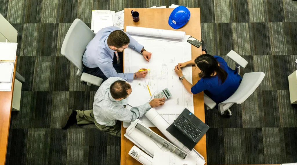 An overhead view of professionals reviewing large architectural blueprints on a desk with a calculator and hard hat, representing architectural review and project oversight services.